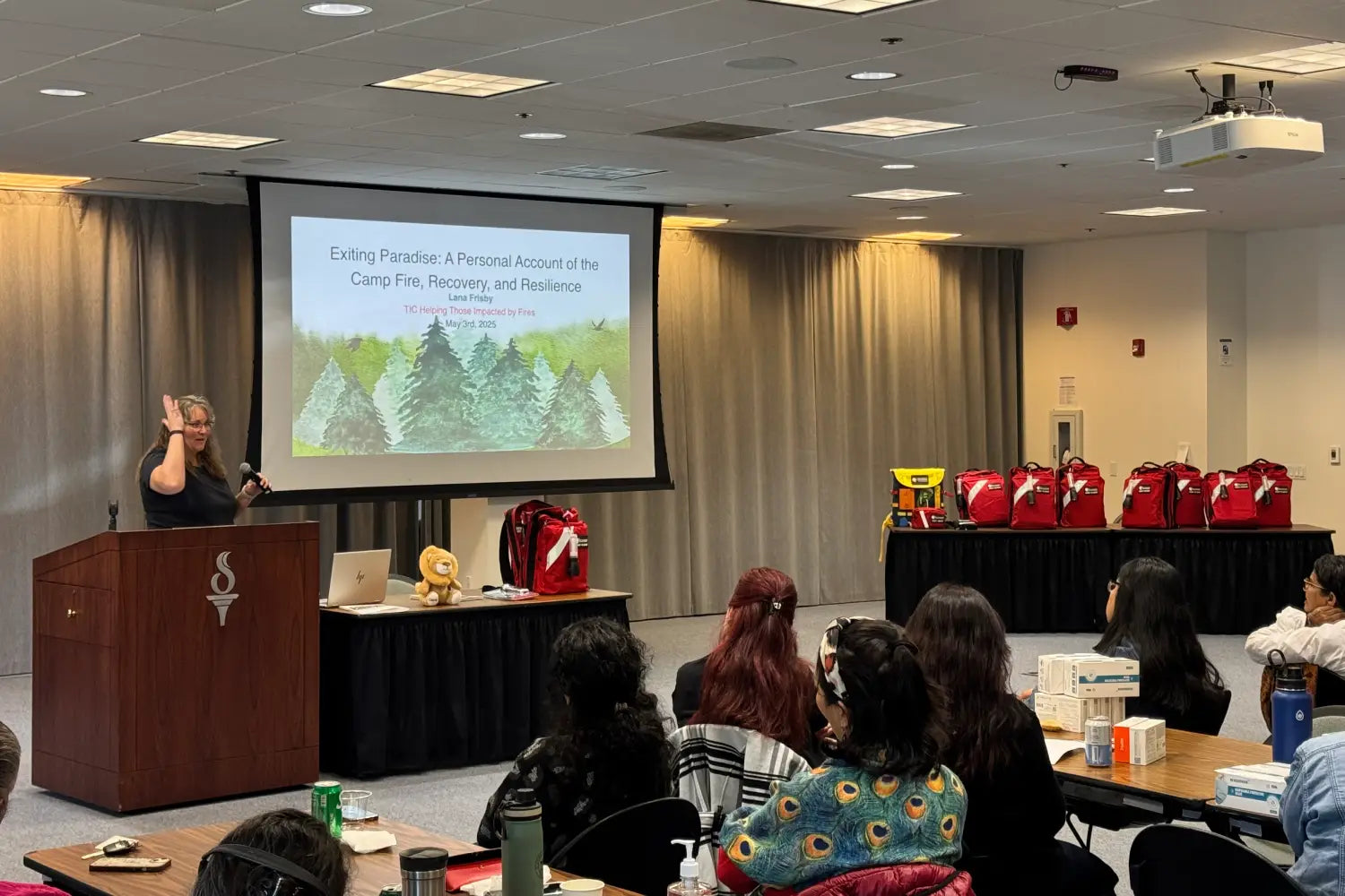Person giving a presentation in a conference room with attendees and a screen displaying a slide.