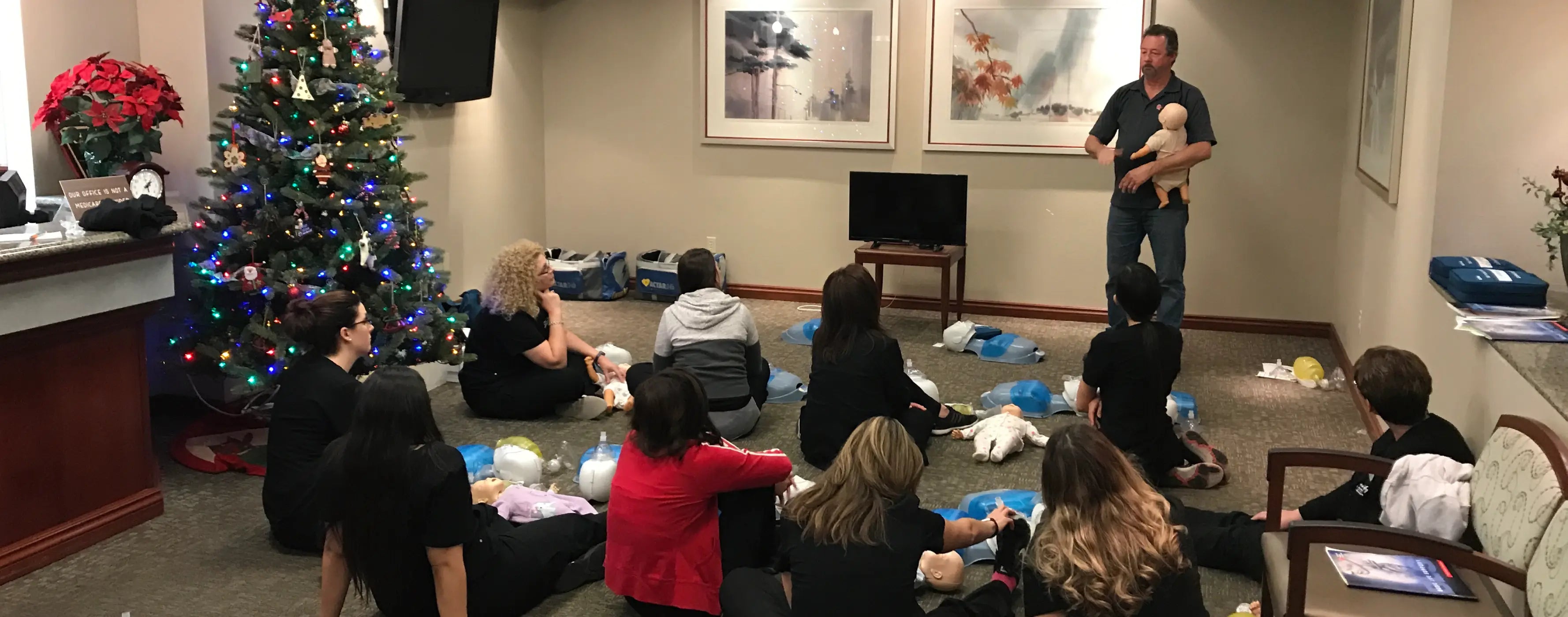 Group of people sitting on the floor in a room with a Christmas tree and a person holding a dog.