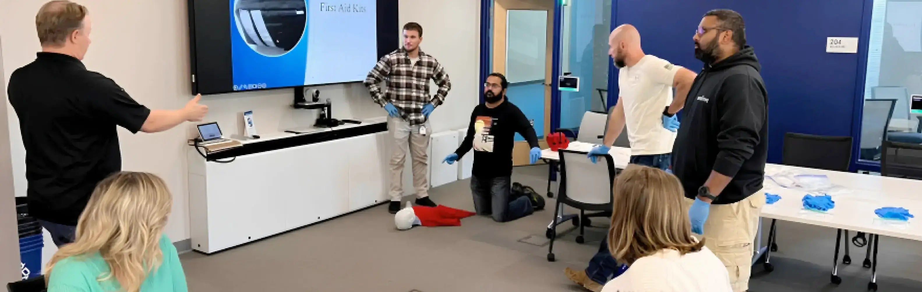 Group of people in a training or meeting room with a presenter using a screen.