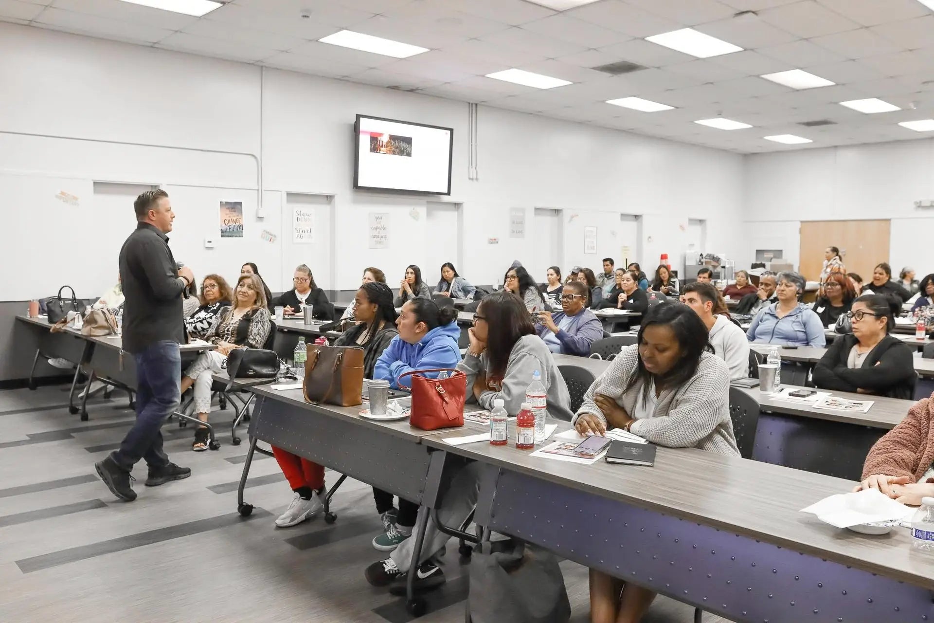 Person speaking to a group of people in a classroom setting with tables and chairs.